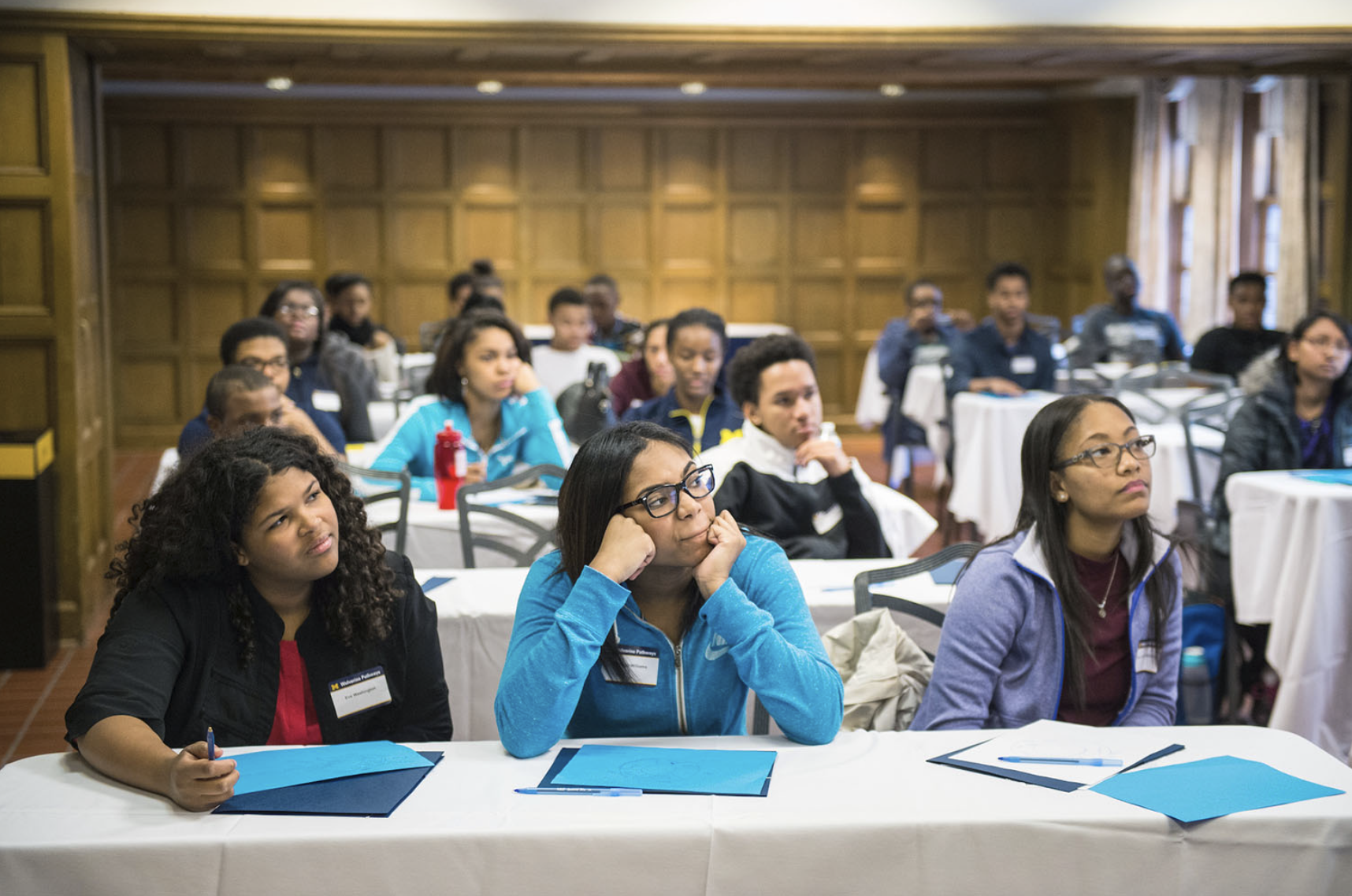 3 female students sitting at a table looking up, with tables of students behind them