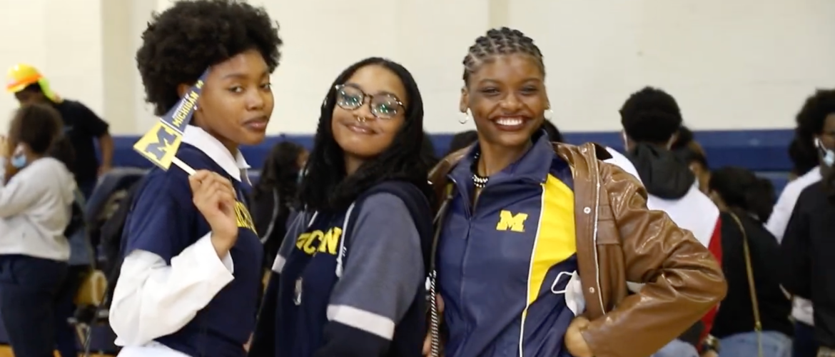 Three people wear Michigan Wolverines apparel, one holding a small Michigan flag. They are indoors, possibly at a sporting event or school gathering.