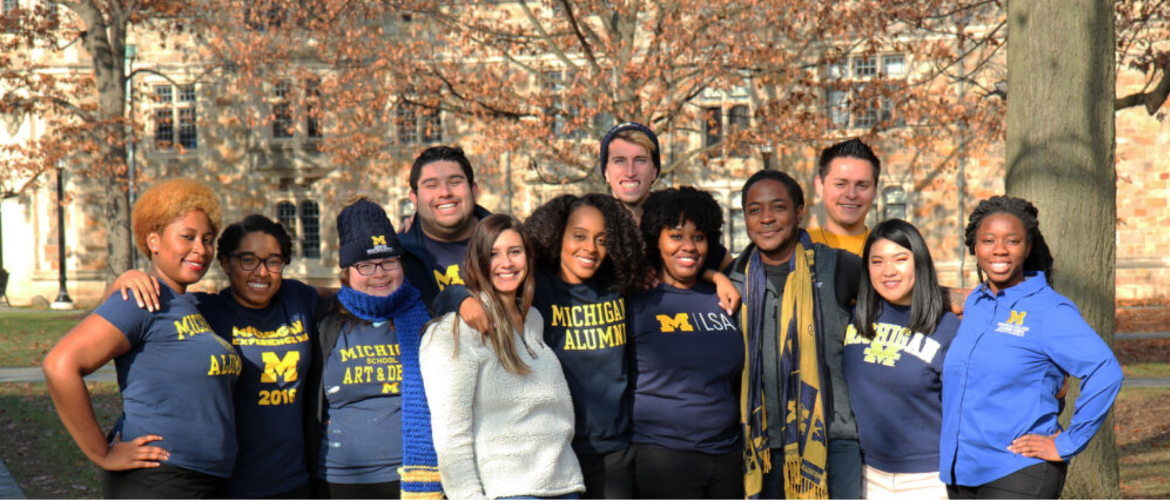 A group of people wearing Michigan Wolverines apparel standing together outside a building with fall foliage in the background.