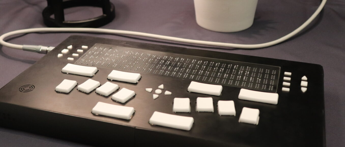 A close-up of a black electronic device with a braille display and white keys, connected by a white cable to an unseen object.