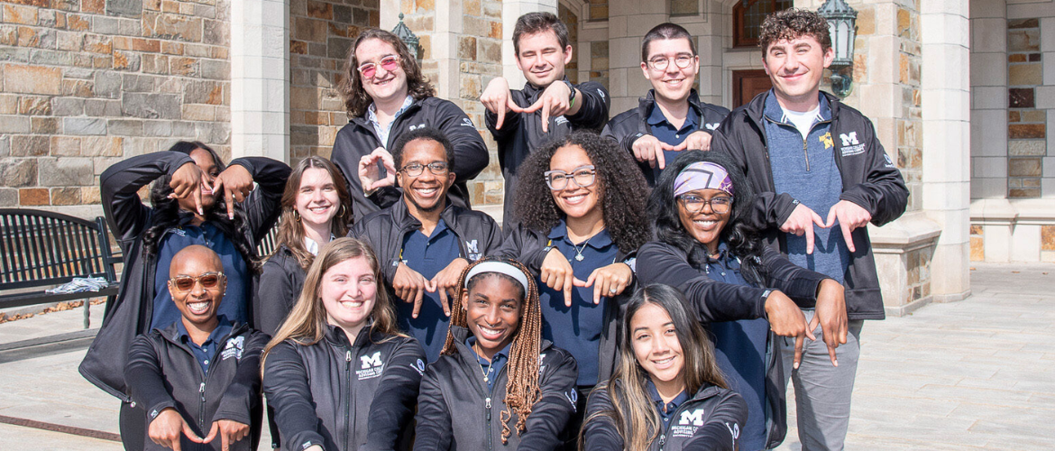 A group of students stand on stone steps in front of a stone building, all posing with their hands in a 