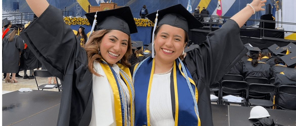 Two graduates in academic regalia with caps and gowns stand in front of a large block M banner, smiling with their arms raised in celebration.