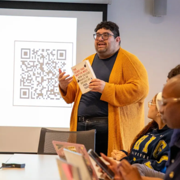 A speaker gestures while holding a book and standing in front of a projection screen displaying 