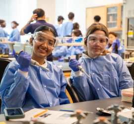 Two individuals in a science lab wearing blue coats and safety goggles, each holding a pipette and smiling at the camera.