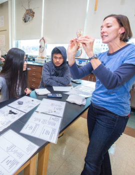 A teacher in a blue shirt holds up a small object while demonstrating to students sitting at desks in a classroom laboratory.