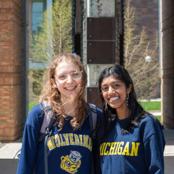 Two smiling individuals wear navy blue sweatshirts with yellow lettering, one reading 