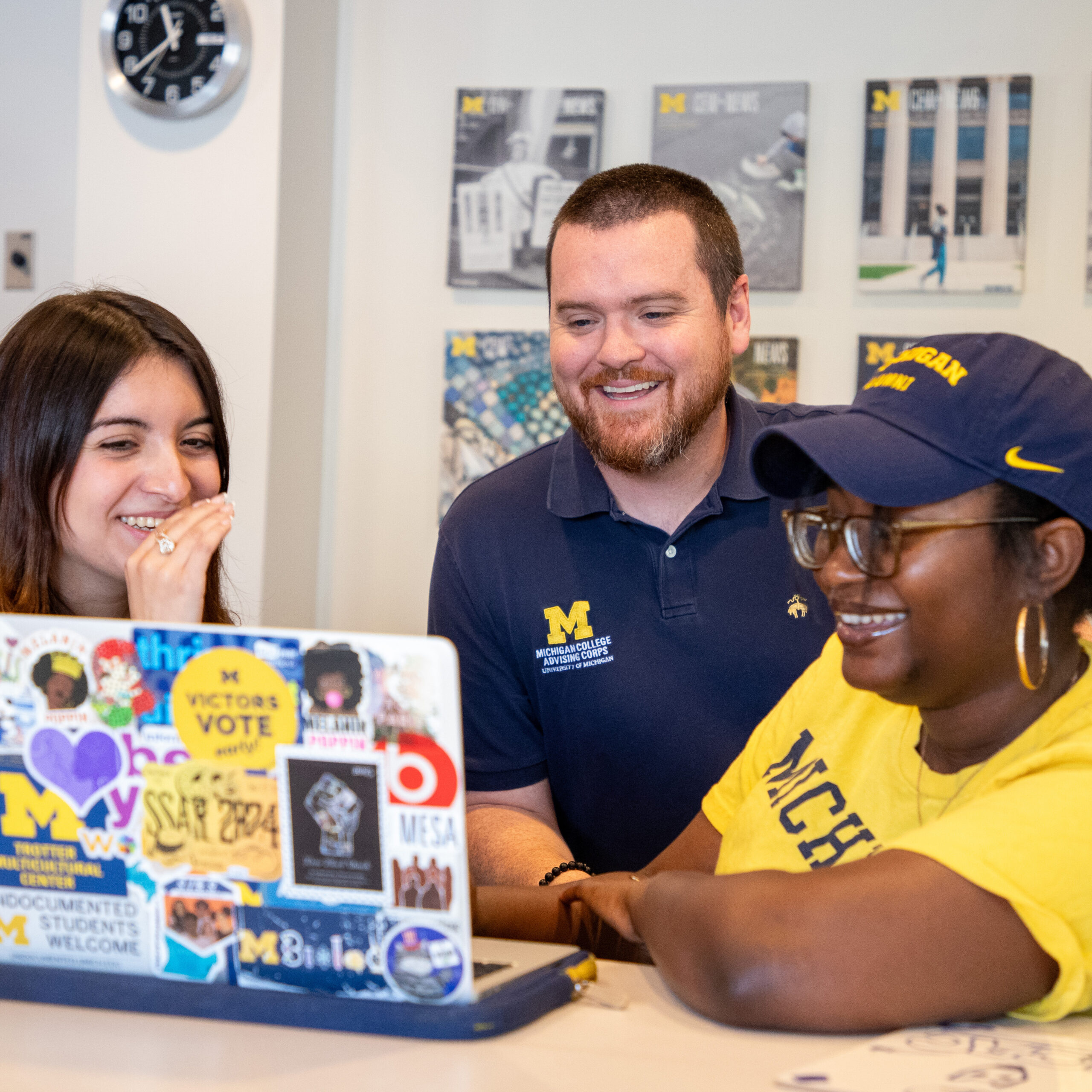 three people sitting looking at a computer screen with lots of stickers on back wearing Michigan clothes and caps.
