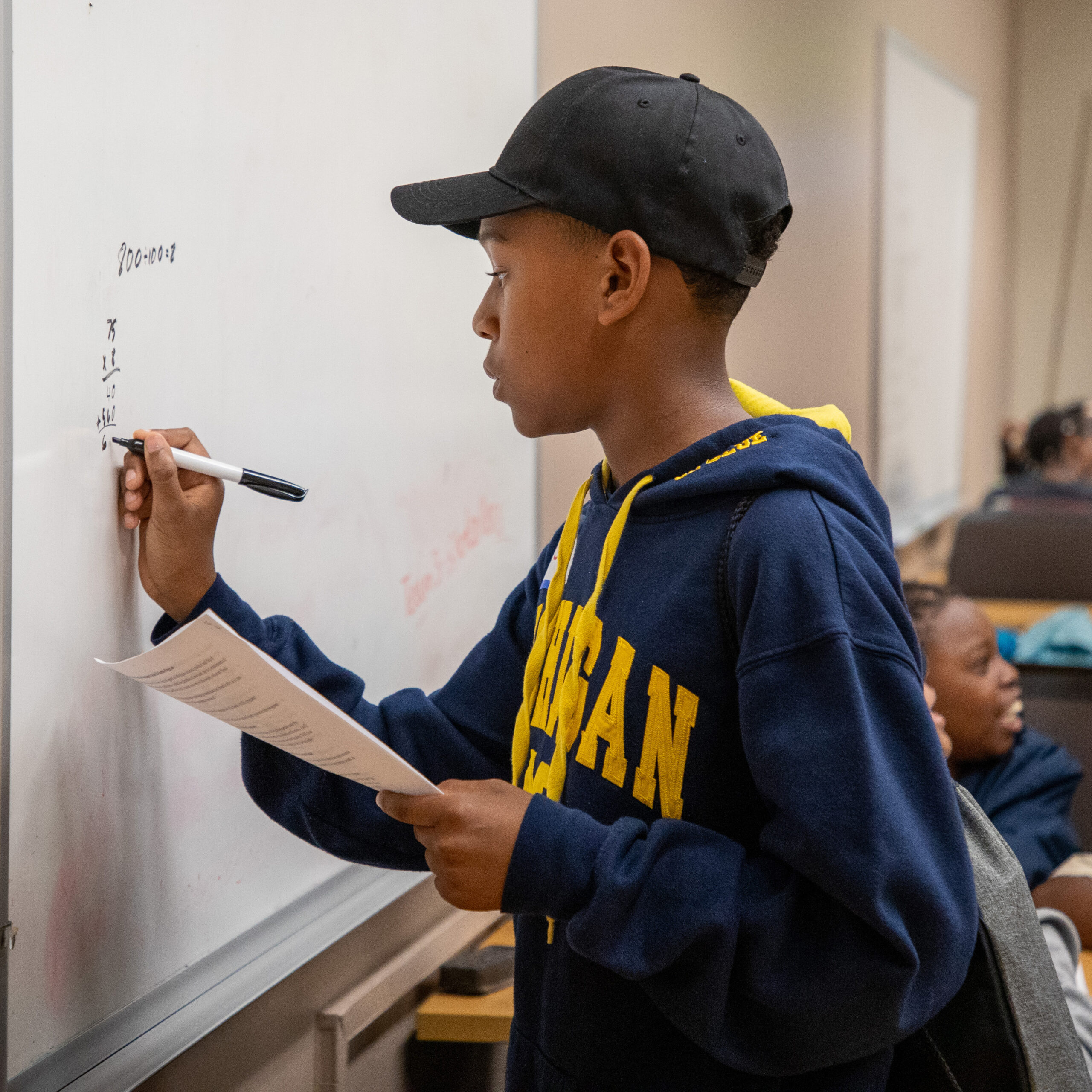 Middle school-age black male wearing a baseball cap and a Michigan sweatshirt writing on a whiteboard