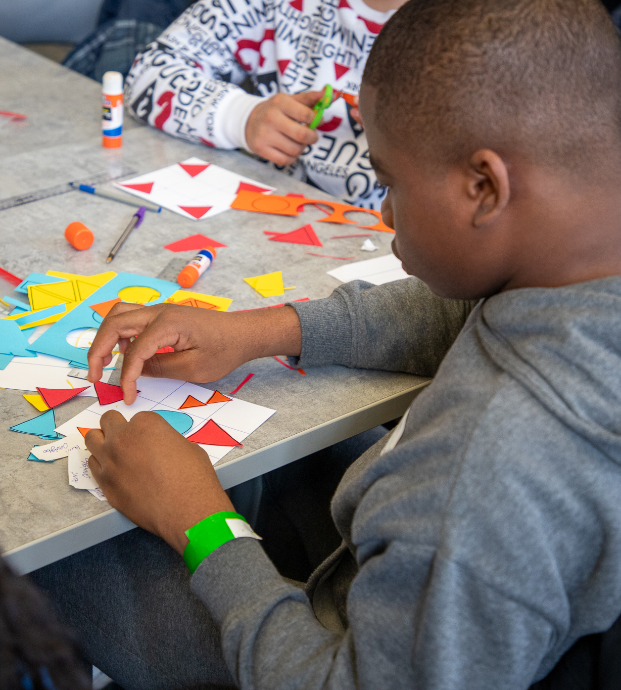 Black male teen wearing grey hoodie sitting at table with cut paper shapes