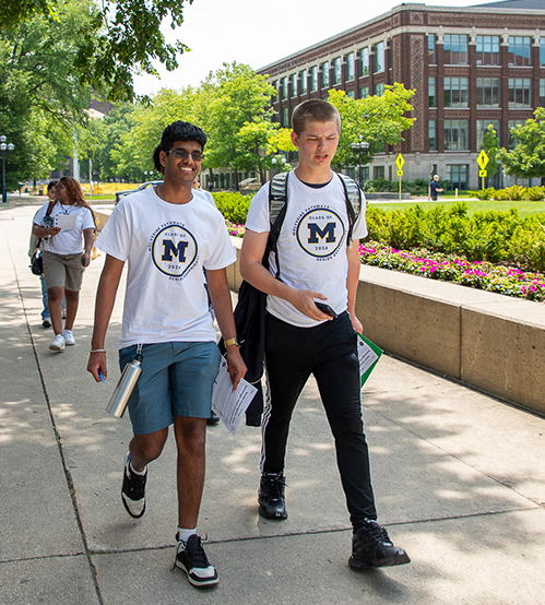 two teenage males, one dark skinned, one light, walking outside on a college campus wearing white t-shirts with navy 