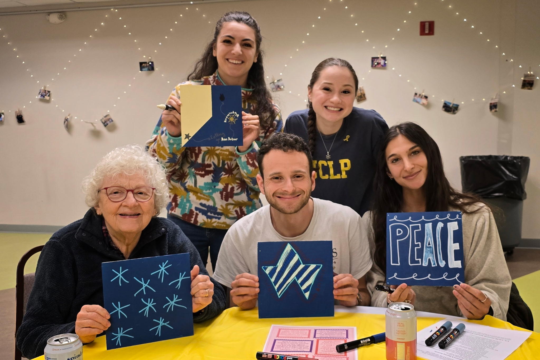 Five smiling people of different generations pose together at a table during a community art activity indoors. Each person holds a hand-painted blue canvas featuring designs and messages such as stars, a ribbon symbol, and the word ‘PEACE,’ with string lights and hanging photos decorating the background.