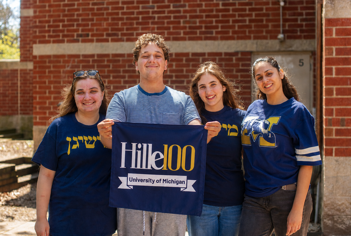Four smiling college students stand outdoors in front of a brick building, holding a ‘Hillel100 University of Michigan’ banner. The group wears University of Michigan-themed blue shirts and poses together in bright sunlight.