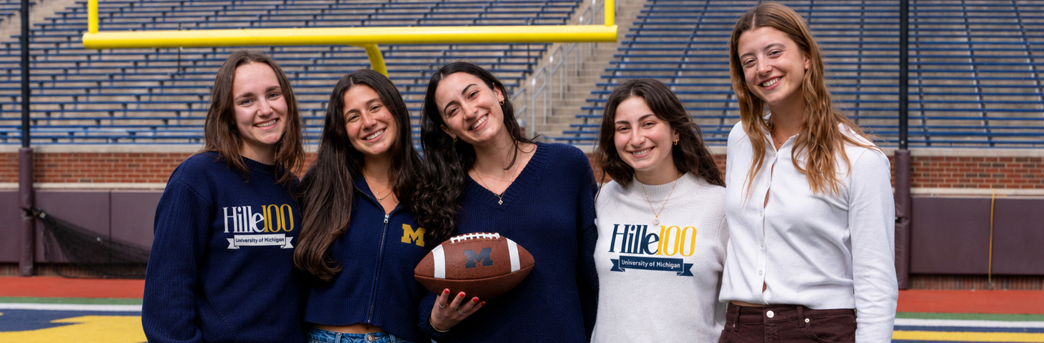 girls standing on football field holding a football wearing Michigan sweatshirts