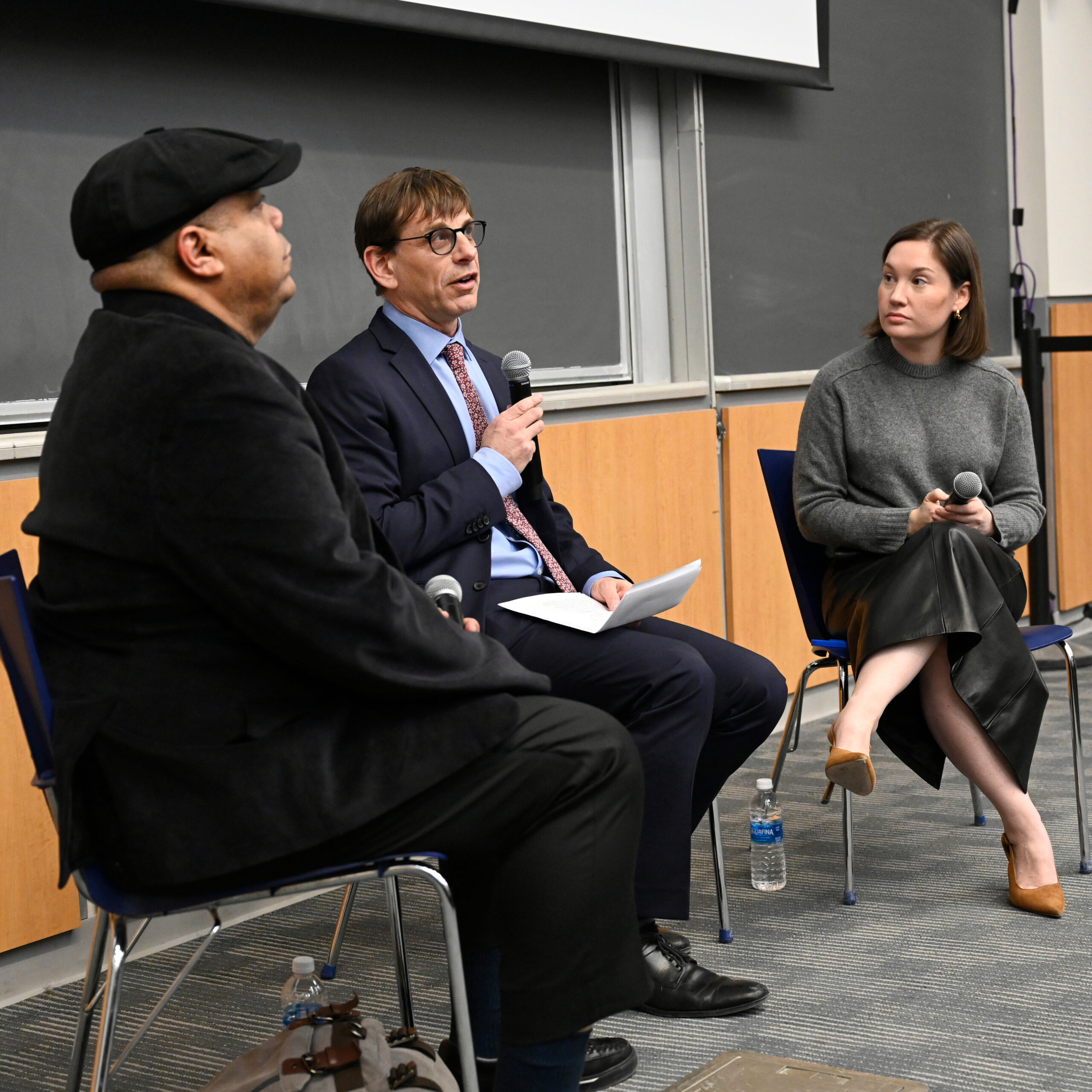 seated from left to right: male in hat, male in tie holding microphone, female turned towards them. all seated