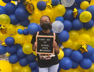 Bethy Carter wearing a mask against a background of maize and blue balloons holding a first day of school sign